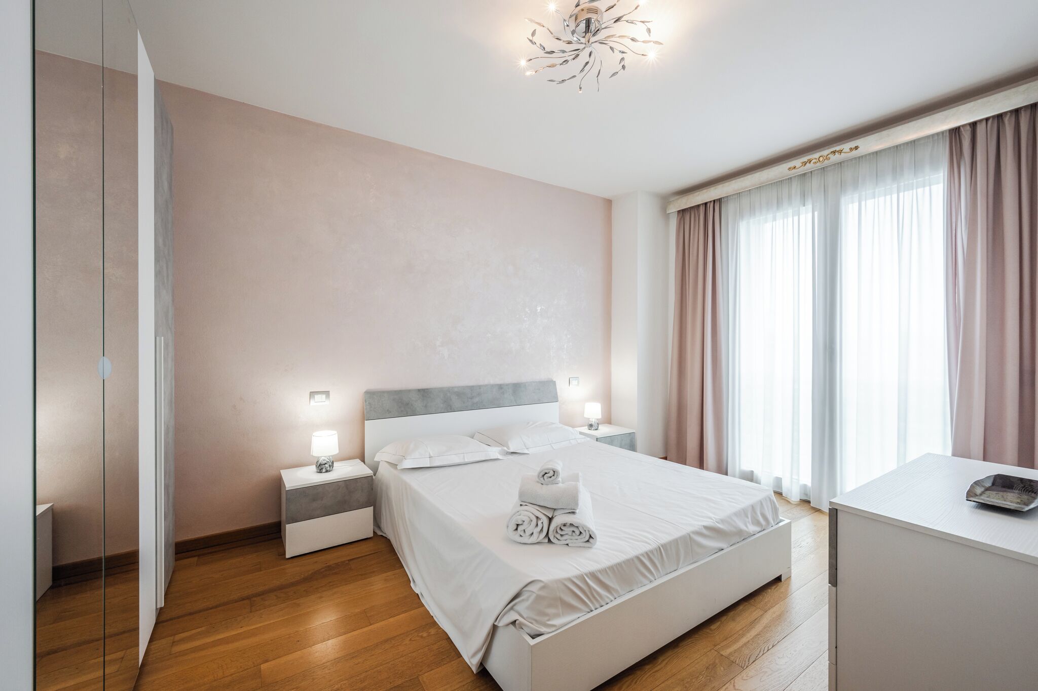 A modern hotel room with a large bed, bedside tables, a desk, and a chandelier.