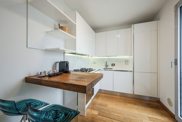 A modern kitchen with white cabinetry, a wooden countertop, and a built-in stove. There are two glass chairs and a small table with decorative items. - Emilia Suite Street (Modena)