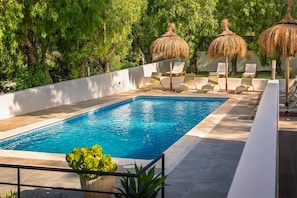 A swimming pool with lounge chairs and thatched umbrellas, surrounded by greenery.
