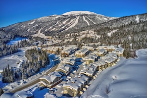 Um resort coberto de neve com vários edifícios em estilo chalé dispostos em fileiras, uma pista de esqui ao fundo e uma paisagem montanhosa.