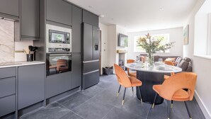 A modern kitchen with grey cabinets, a built-in oven, and a dining area with a round table and orange chairs. - Birdsong Cottage - North East Escapes (Belford)
