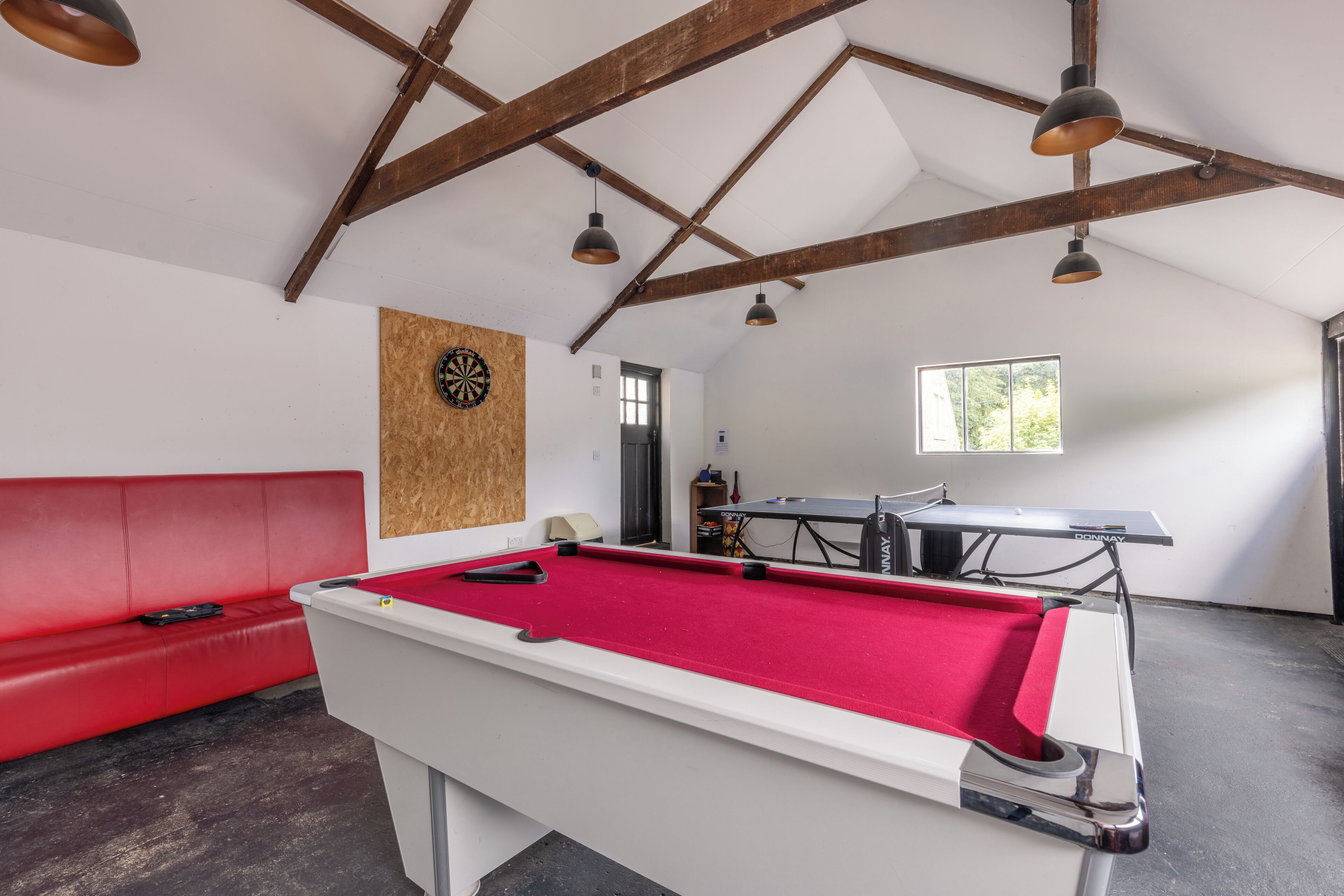 Une table de billard recouverte d’un drap rouge, un canapé rouge et des poutres en bois au plafond.