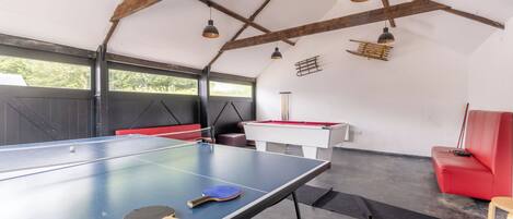 A ping pong table with two paddles, a red sofa, and a pool table in a room with exposed wooden beams and large windows.