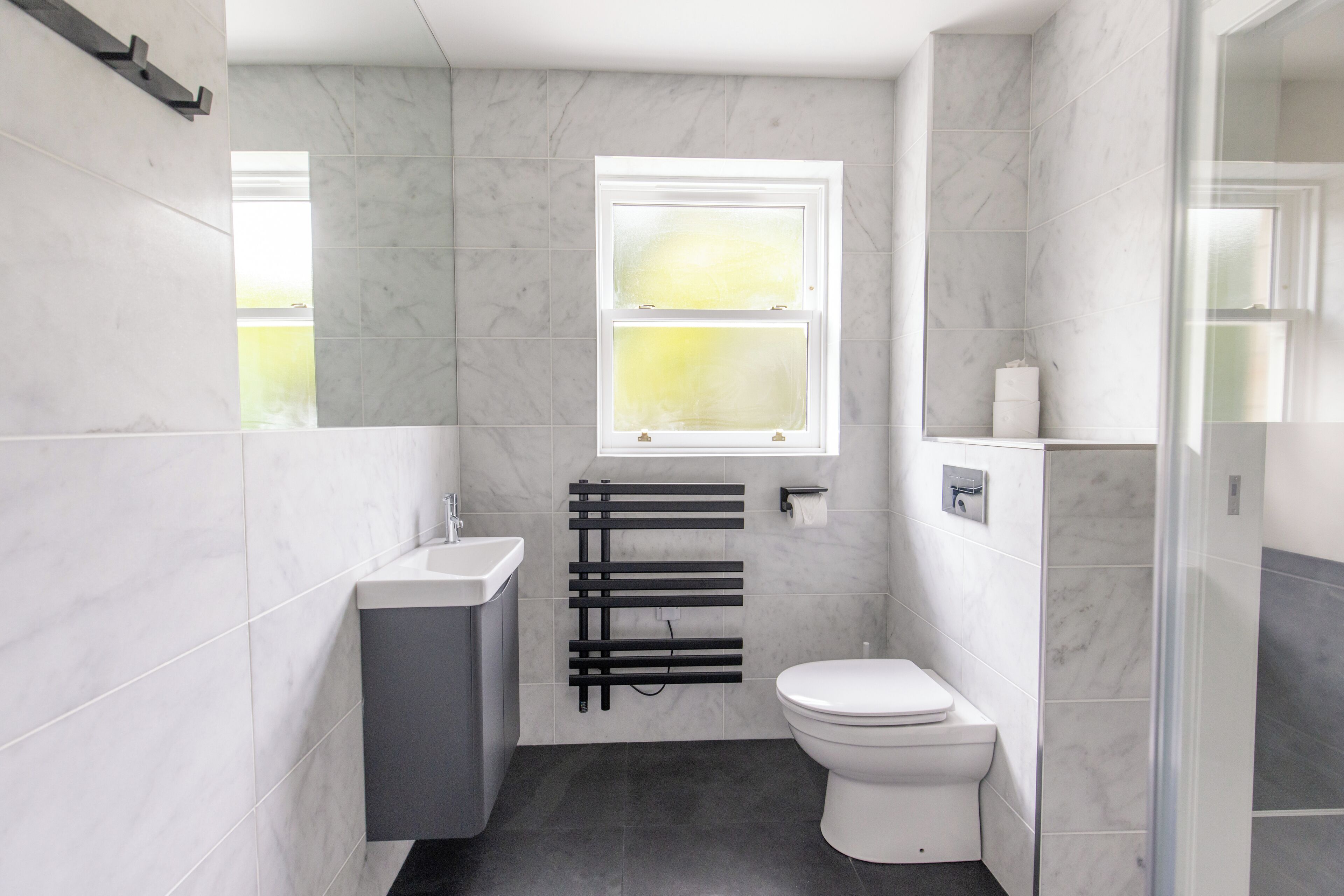 Une salle de bain moderne avec des toilettes blanches, un porte-serviettes noir et une fenêtre laissant entrer la lumière naturelle.