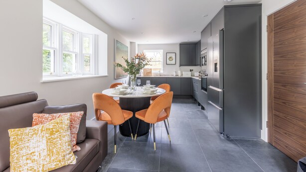 A modern kitchen with dark cabinets, a central island, and a dining area with a round table and orange chairs. - Birdsong Cottage - North East Escapes (Belford)