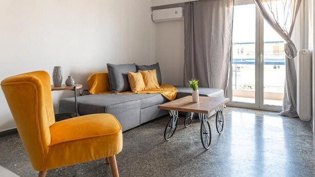 Ein modernes Wohnzimmer mit einer Couch, einem gelben Sessel, einem Couchtisch und Blick ins Freie durch ein großes Fenster.