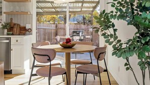 Un coin repas avec une table ronde, quatre chaises et un saladier de fruits. Le sol est en bois, il y a un luminaire au plafond et, grâce à une porte vitrée, on peut admirer la vue sur l’extérieur.