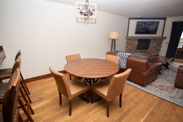 A dining area with a round wooden table and six chairs, a stone fireplace, a sofa, and a lamp. - Ski-In / Ski-Out! Hot Tub! New Listing! Garage! (Mt. Crested Butte)
