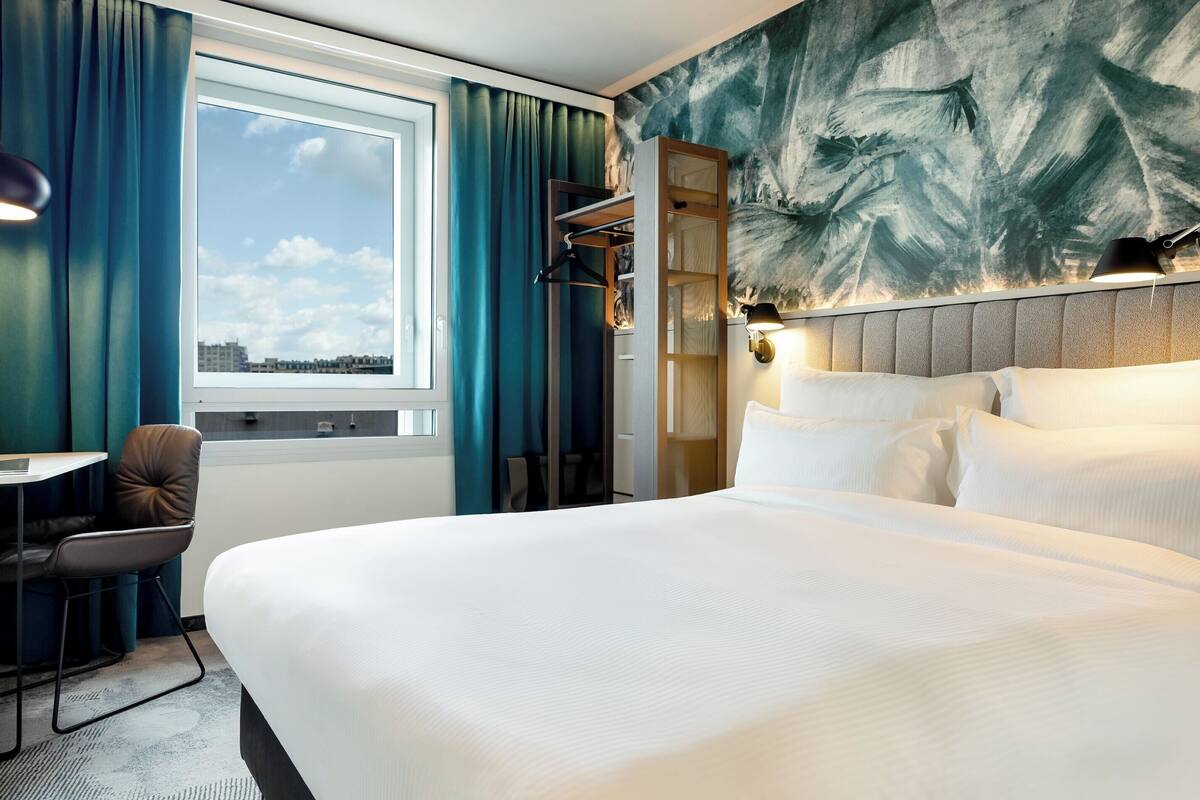 Habitación de hotel con una cama grande, un escritorio y una silla. Tiene una ventana con cortinas, una lámpara de pared y un cuadro de palmeras.