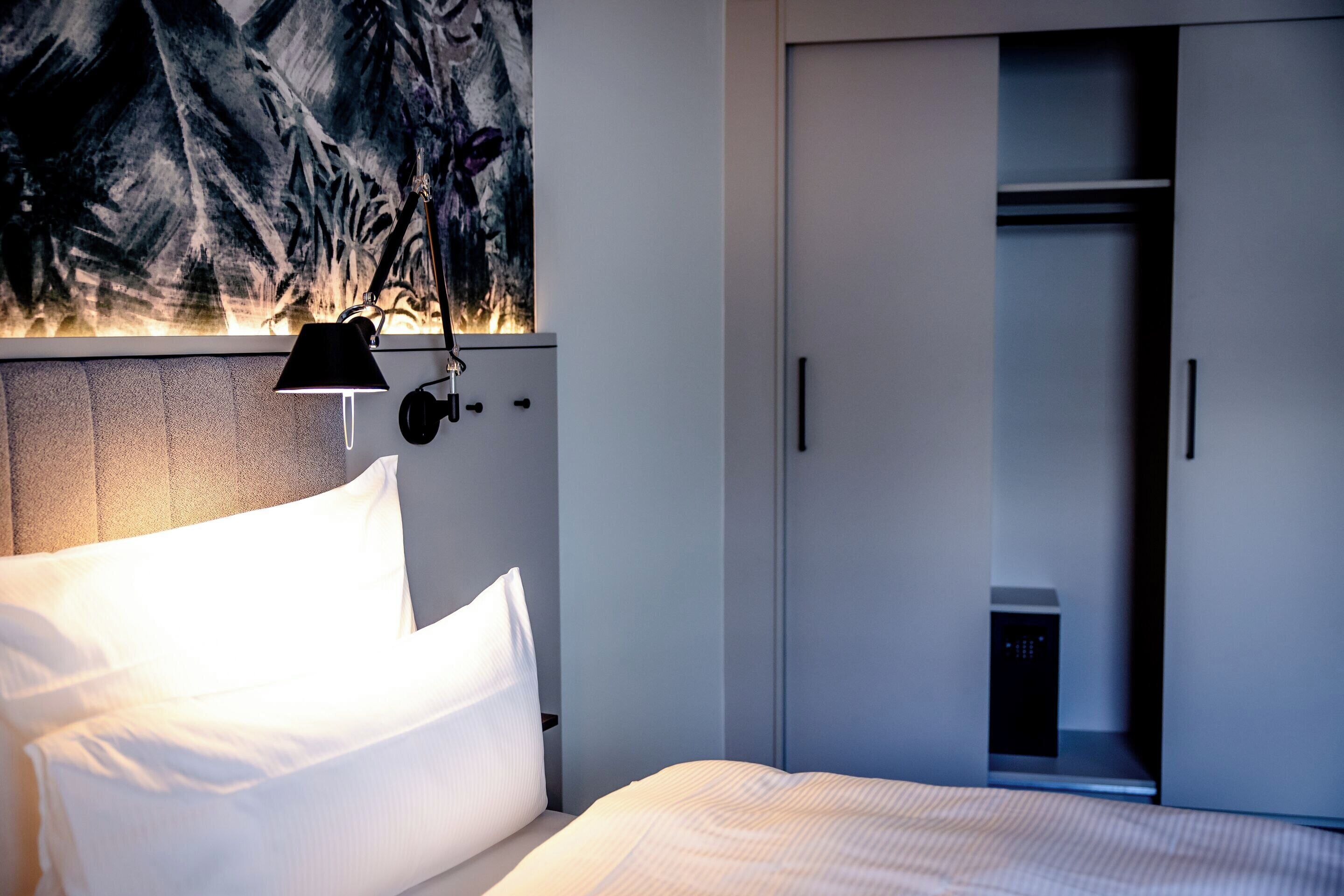 A hotel room with a bed, a nightstand, a wall-mounted lamp, and a wardrobe.