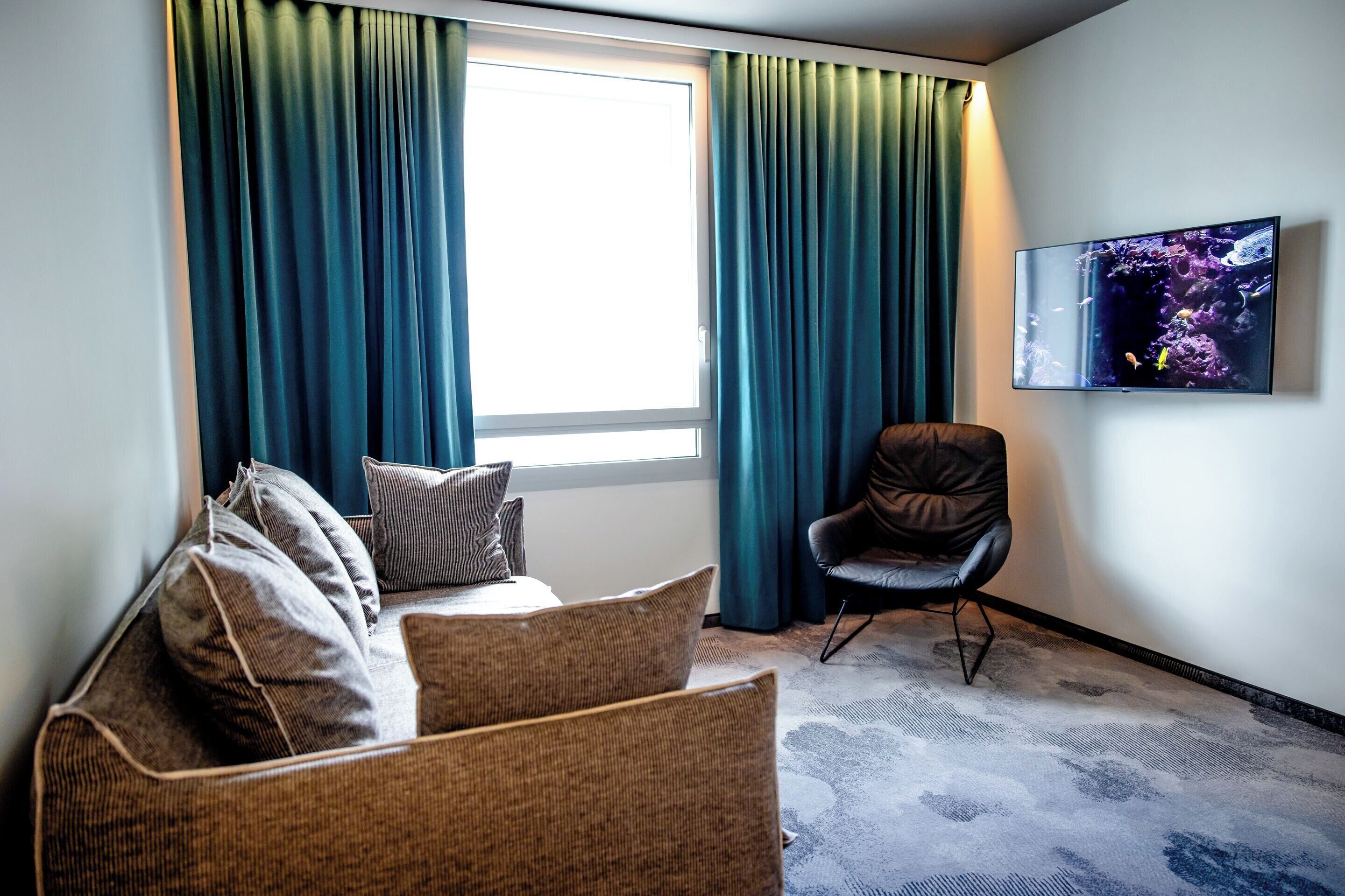 Ein Zimmer mit einer Couch, einem Sessel, einem großen Fenster mit Vorhängen und einem an der Wand befestigten Fernseher, der eine Unterwasserwelt zeigt.
