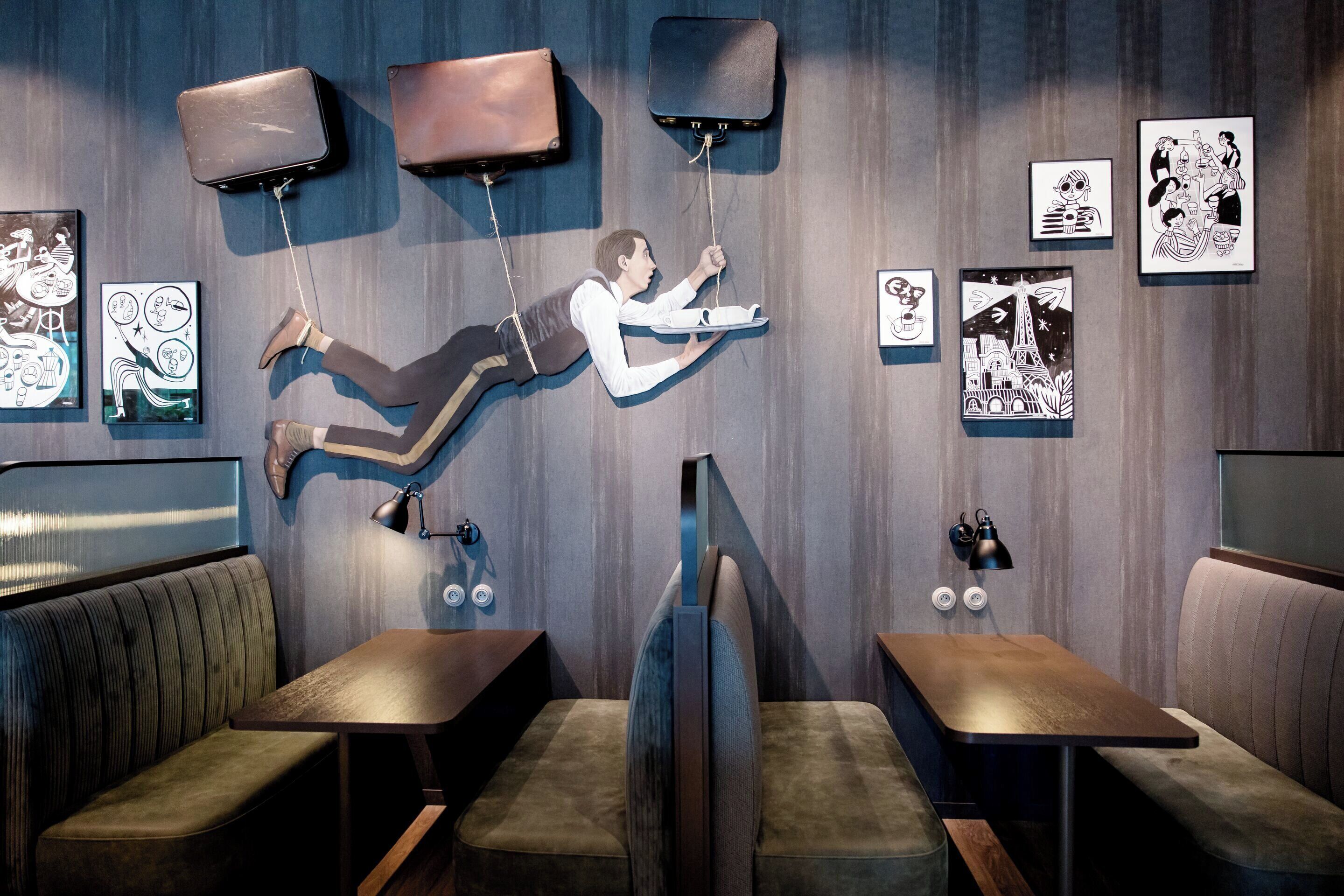 A modern interior with a unique art installation featuring a figure suspended by ropes. There are cushioned seating areas and framed artworks on the walls.