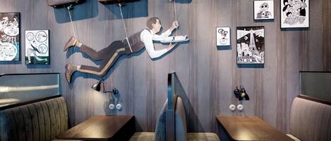 A modern interior with a unique art installation featuring a figure suspended by ropes. There are cushioned seating areas and framed artworks on the walls.