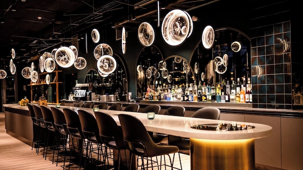 A modern bar area with a long counter, black chairs, and circular hanging lights. - Motel One Paris - Porte de Versailles (Issy-les-Moulineaux)