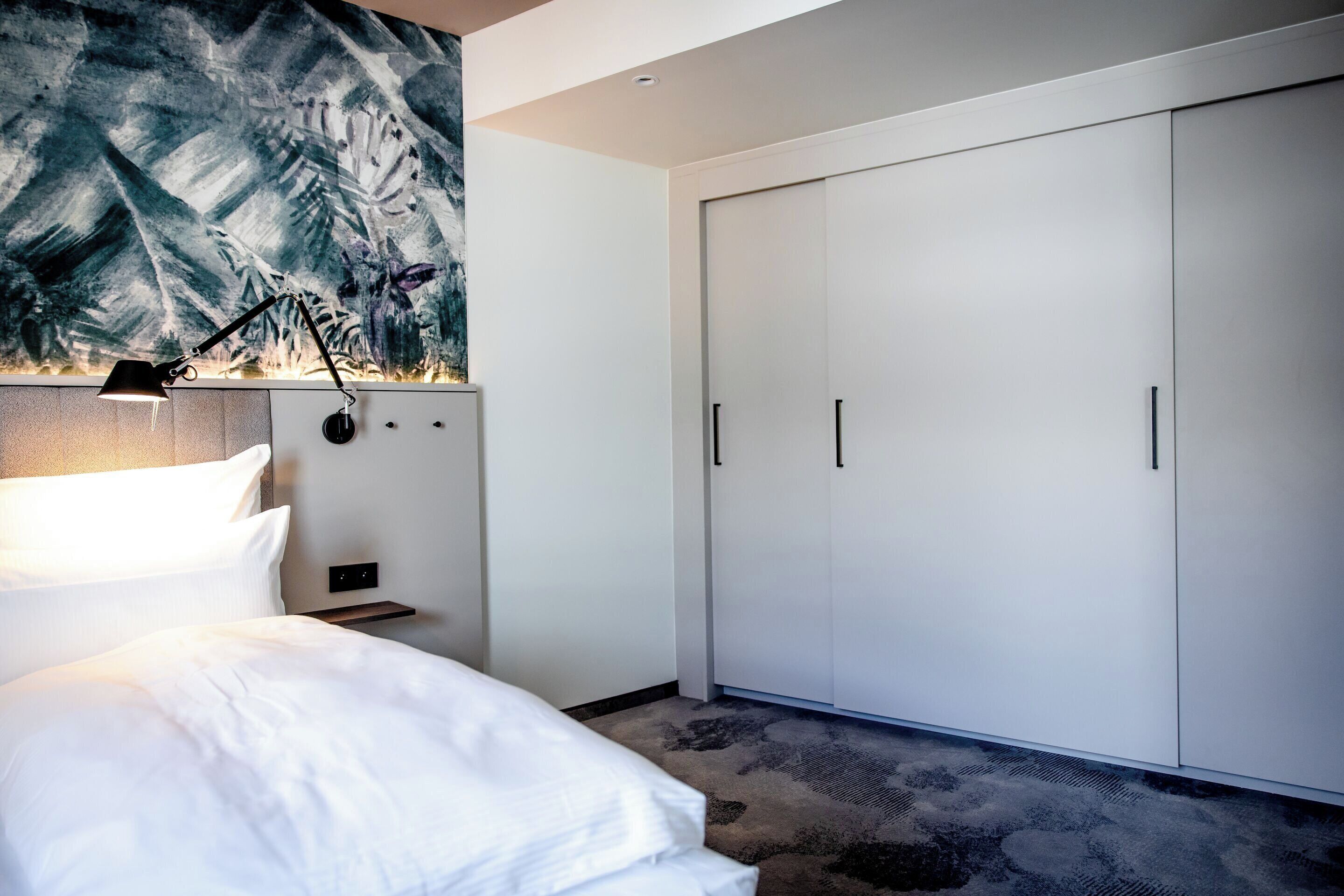 A modern hotel room with a large bed, a nightstand with a lamp, a wardrobe, and a wall-mounted artwork.
