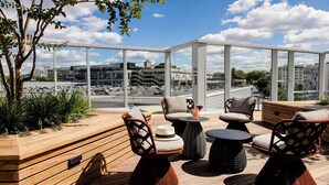 A rooftop terrace with wicker furniture, a wooden bench, and a view of the cityscape. - Motel One Paris - Porte de Versailles (Issy-les-Moulineaux)