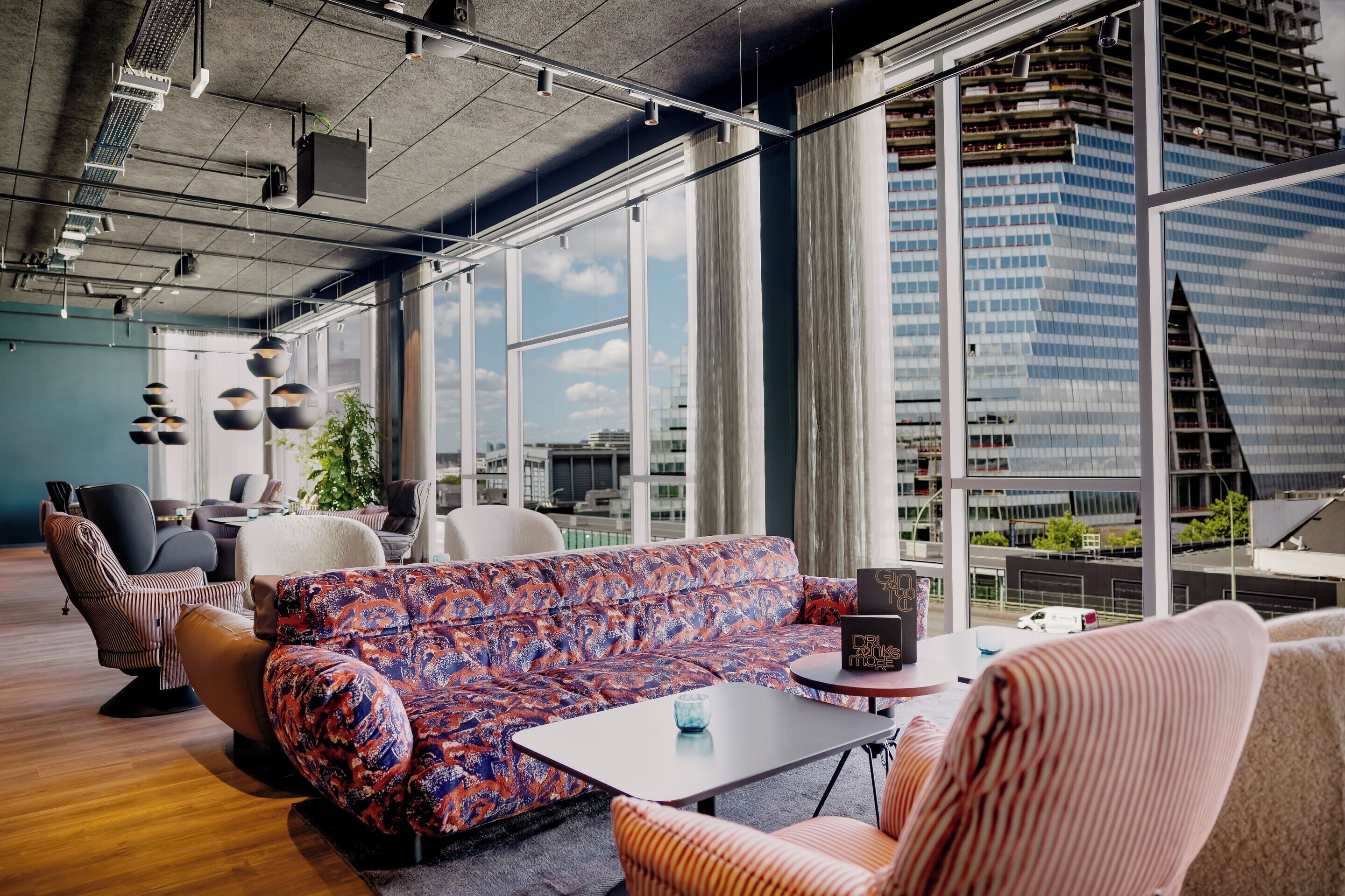 Un salón moderno con un sofá de colores, sillas a rayas y vistas a un paisaje urbano a través de amplios ventanales.