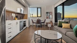 A modern apartment with a kitchen, dining area, and living room. It features a large window with a city view, a dining table with chairs, and a sofa with cushions. - Tower Views! Pool, FREE Banff Pass! (Calgary)