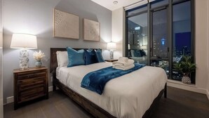 A modern bedroom with a large bed, two bedside tables, and a city view through floor-to-ceiling windows. - Tower Views! Pool, FREE Banff Pass! (Calgary)