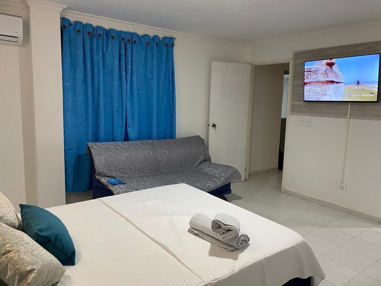 A hotel room with a bed, a grey sofa, a blue curtain, and a TV on the wall.
