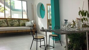 Ein modernes Wohnzimmer mit einer Couch, zwei Sesseln, einem Couchtisch und einem Esstisch mit Stühlen. Ein großes Fenster, eine türkisfarbene Akzentwand und eine Pflanze in der Ecke sind zu sehen.