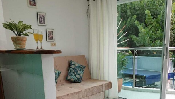 Uma sala de estar moderna com sofá, mesa de centro de vidro e uma área de jantar com mesa e cadeiras. Há uma porta de vidro deslizante que dá para uma varanda com piscina.