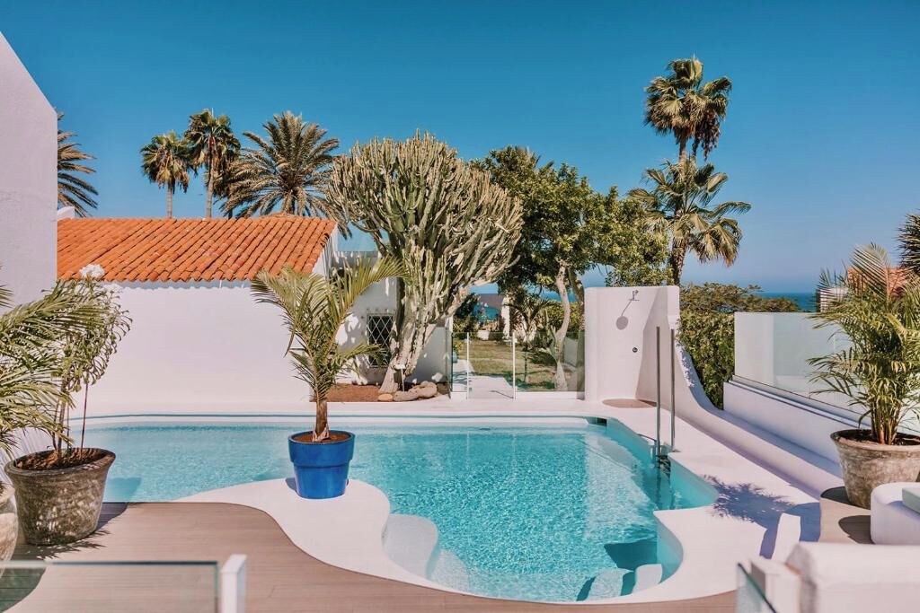 A pool area with a clear view of the ocean, surrounded by palm trees and a modern building with a red-tiled roof.