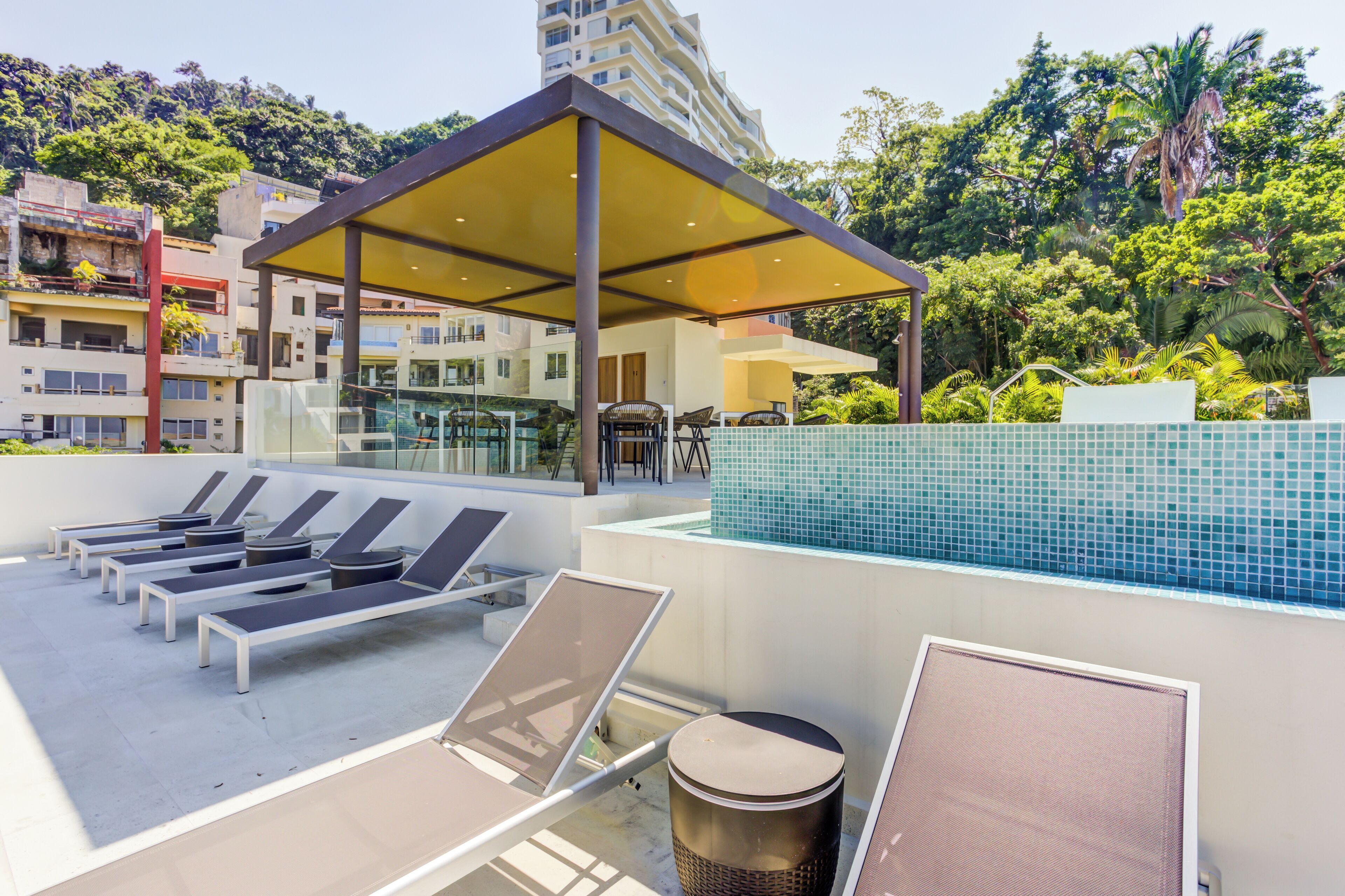 A rooftop pool area with sun loungers, a covered seating area, and a view of surrounding buildings and greenery.