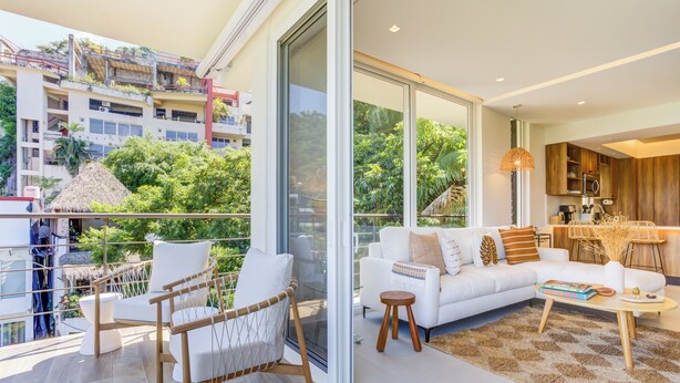 Un salon moderne avec un canapé blanc, une table basse en bois et une porte vitrée donnant sur un balcon offrant une vue sur le paysage urbain et la verdure.