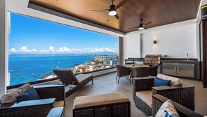 A balcony with wicker furniture, a grill, and a view of the sea and buildings.