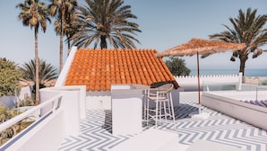 Una terraza en la azotea con techo de tejas, una mesa con sillas y un parasol de paja.