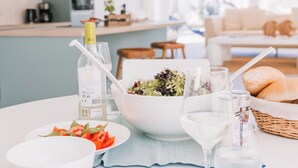 Mesa de comedor dispuesta con platos, tazones y vasos blancos, una botella de vino blanco y una cesta de pan.