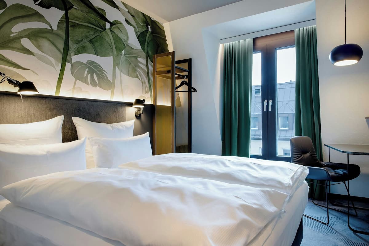A hotel room with a large bed, a desk with a chair, a mirror, and a window with curtains.
