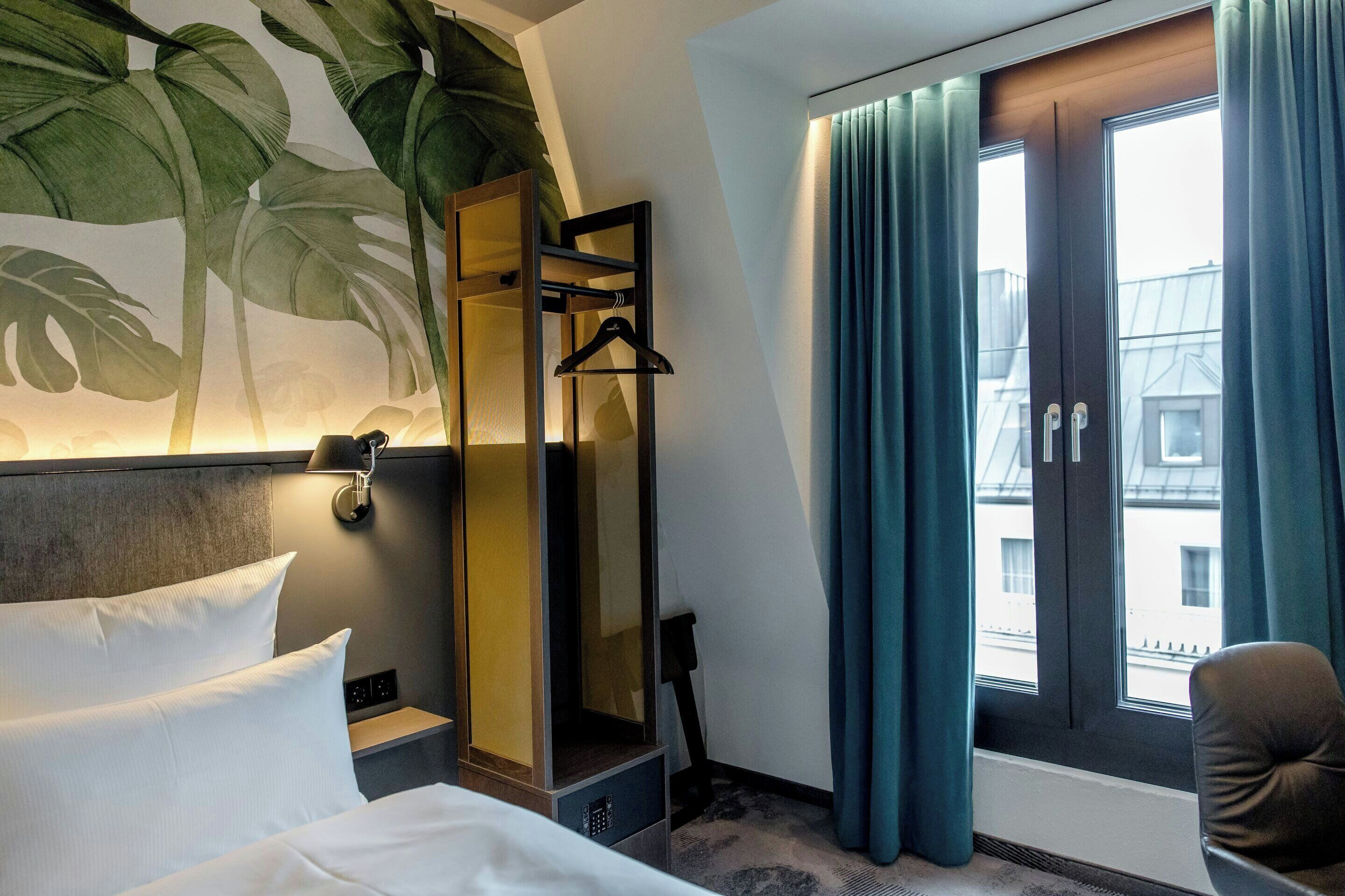 A hotel room with a bed, a mirror, a hanging coat rack, a chair, and a window with curtains.