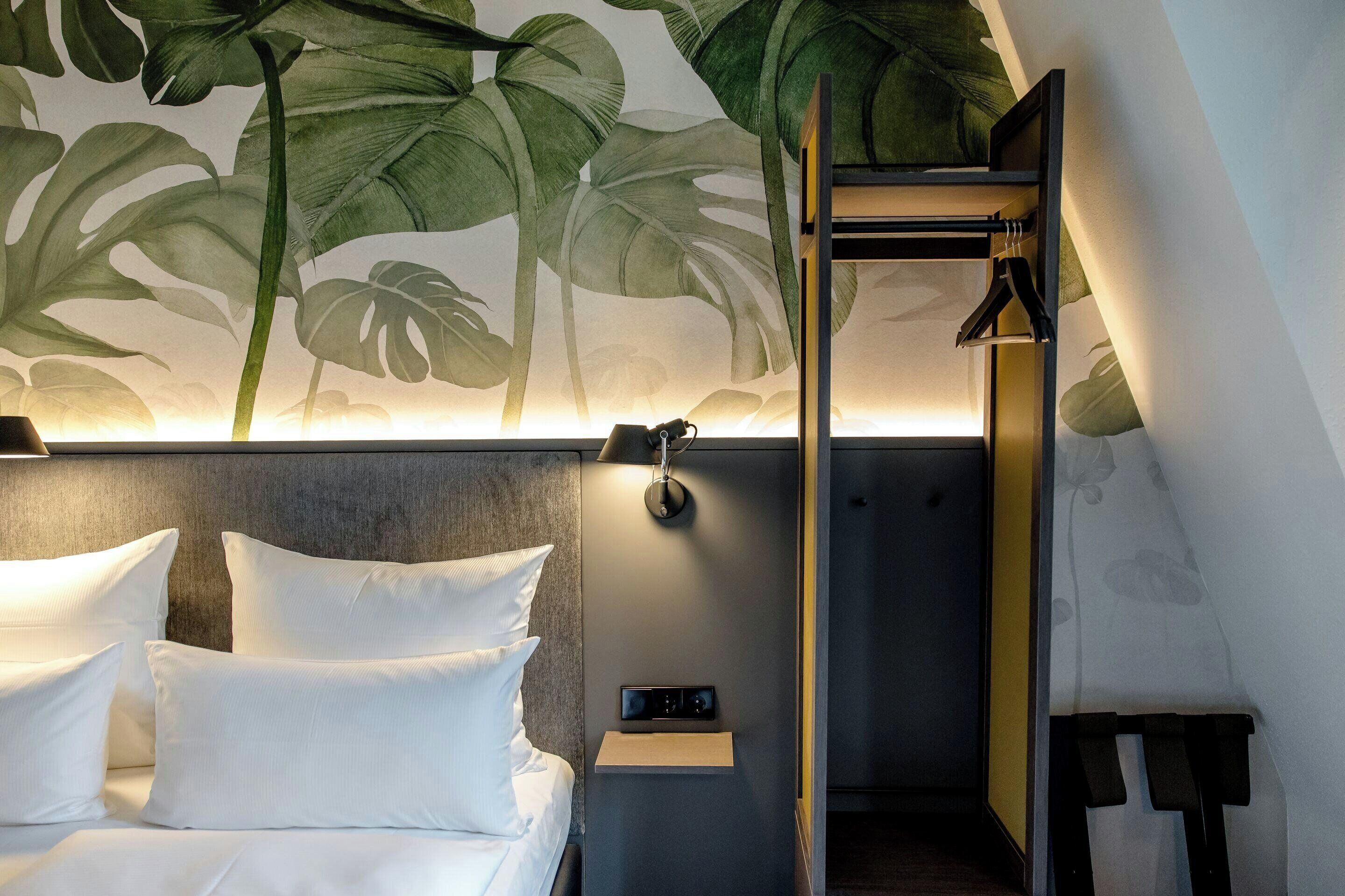 Habitación de hotel con cama, mesita de noche, luz de pared y un mural de hojas tropicales.
