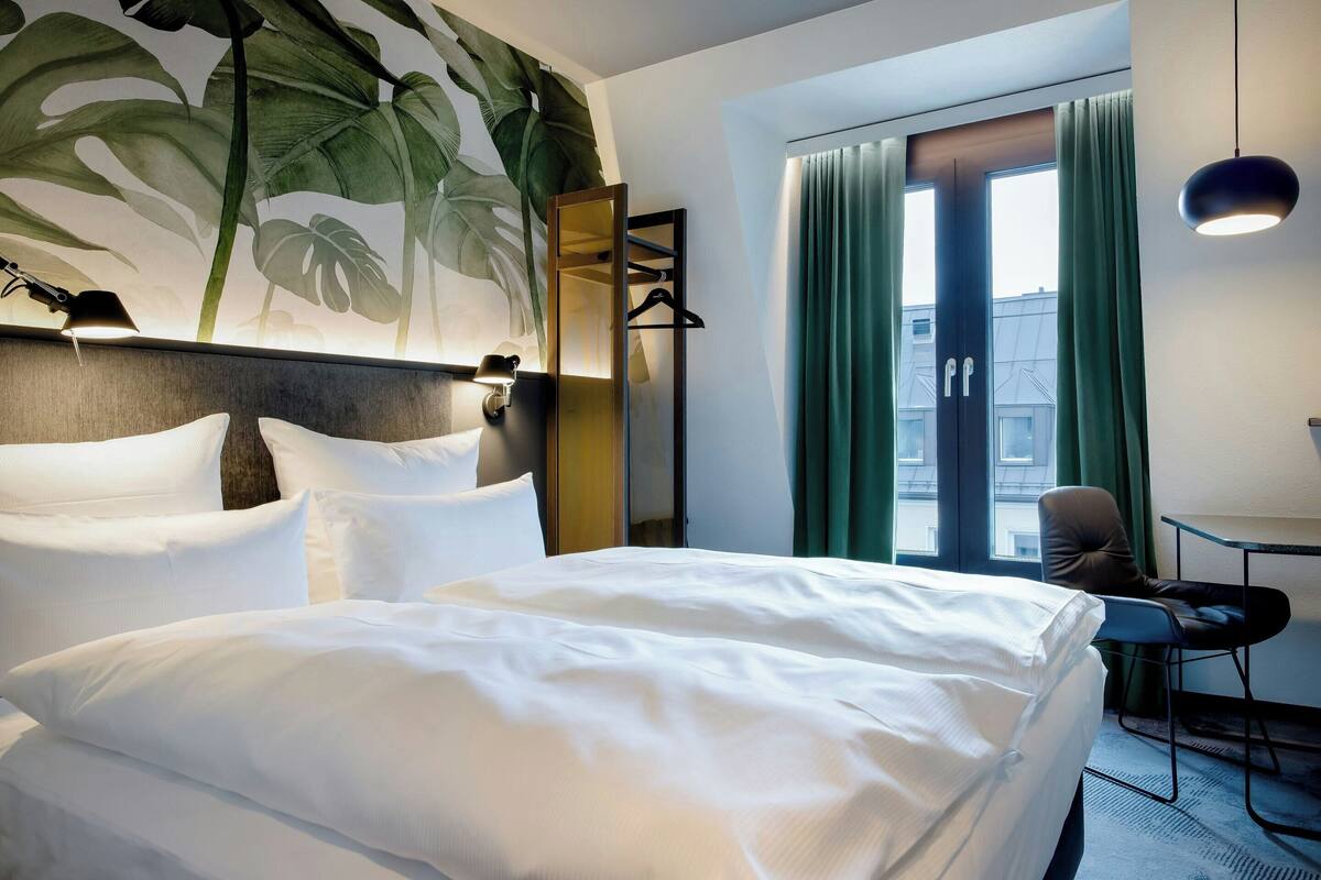 Quarto de hotel com uma cama grande, uma escrivaninha com cadeira, um espelho e uma janela com cortinas.
