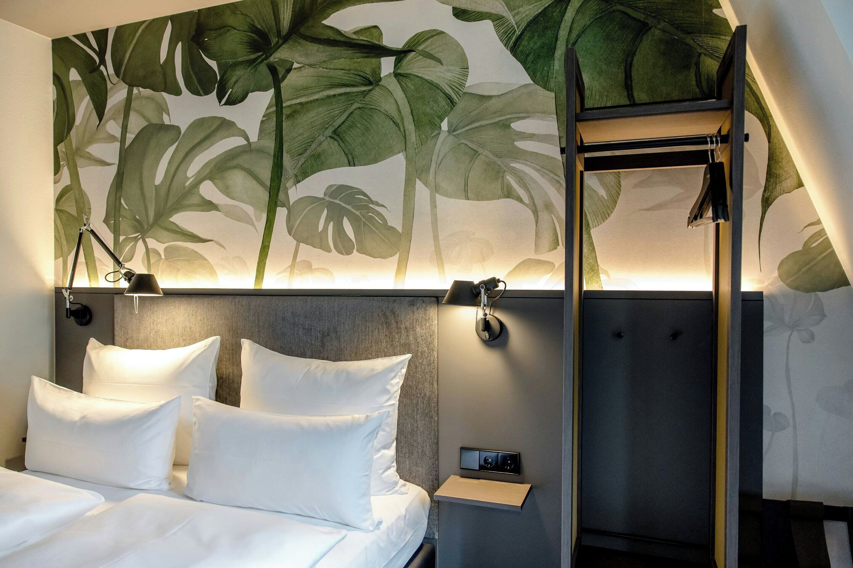 Ein Hotelzimmer mit Bett, weißen Kissen, einem Nachttisch mit Lampe und einer Wand mit tropischem Blatt-Mural.