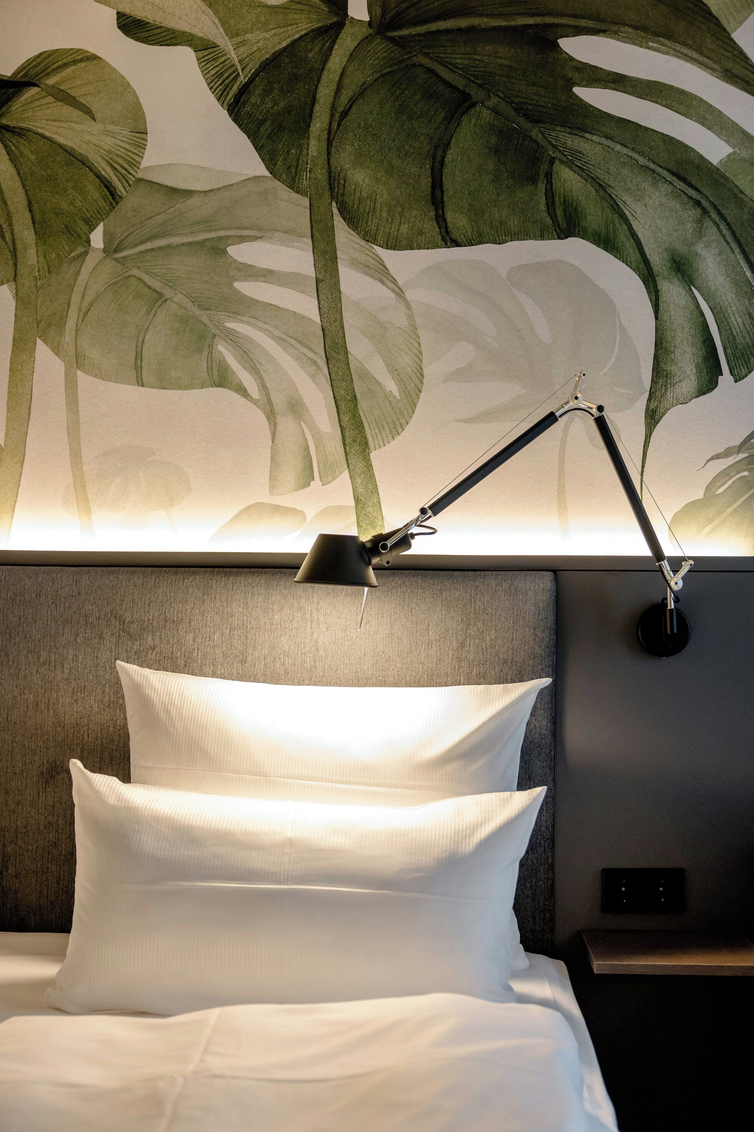Habitación de hotel con cama, almohadas blancas, mesita de noche, lámpara y un mural vegetal en la pared.
