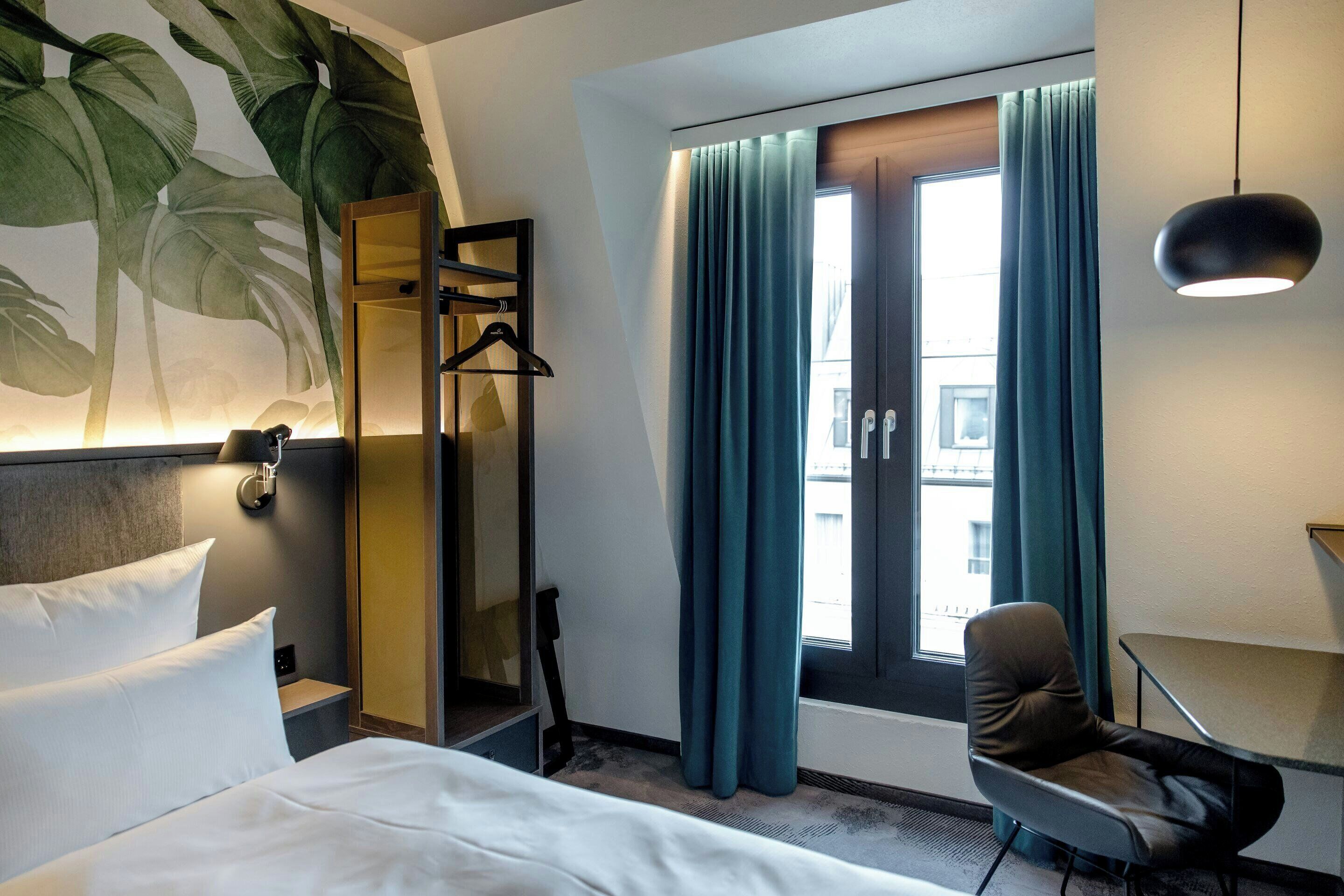 A hotel room with a bed, a chair, a desk, a mirror, and a window with curtains.