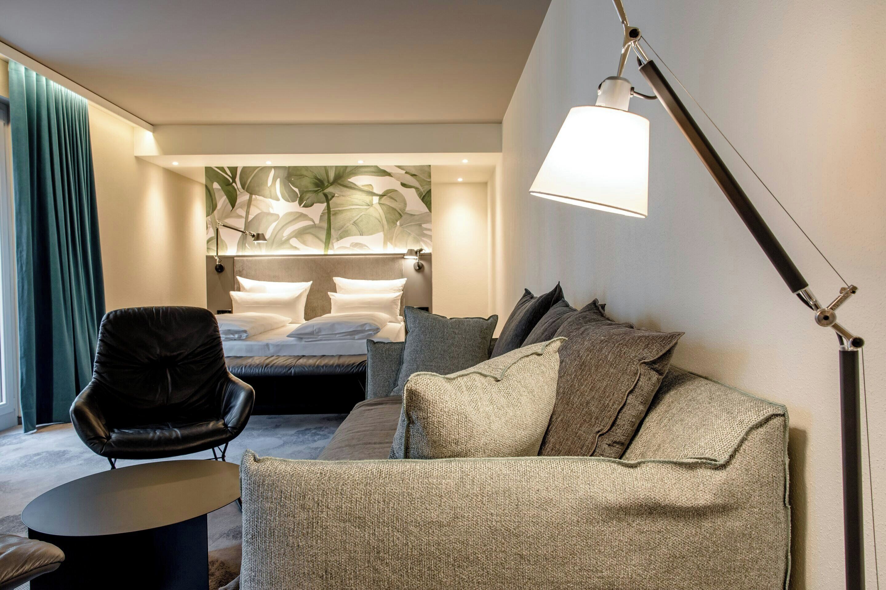 A modern hotel room with a bed, a sofa, a chair, and a lamp.