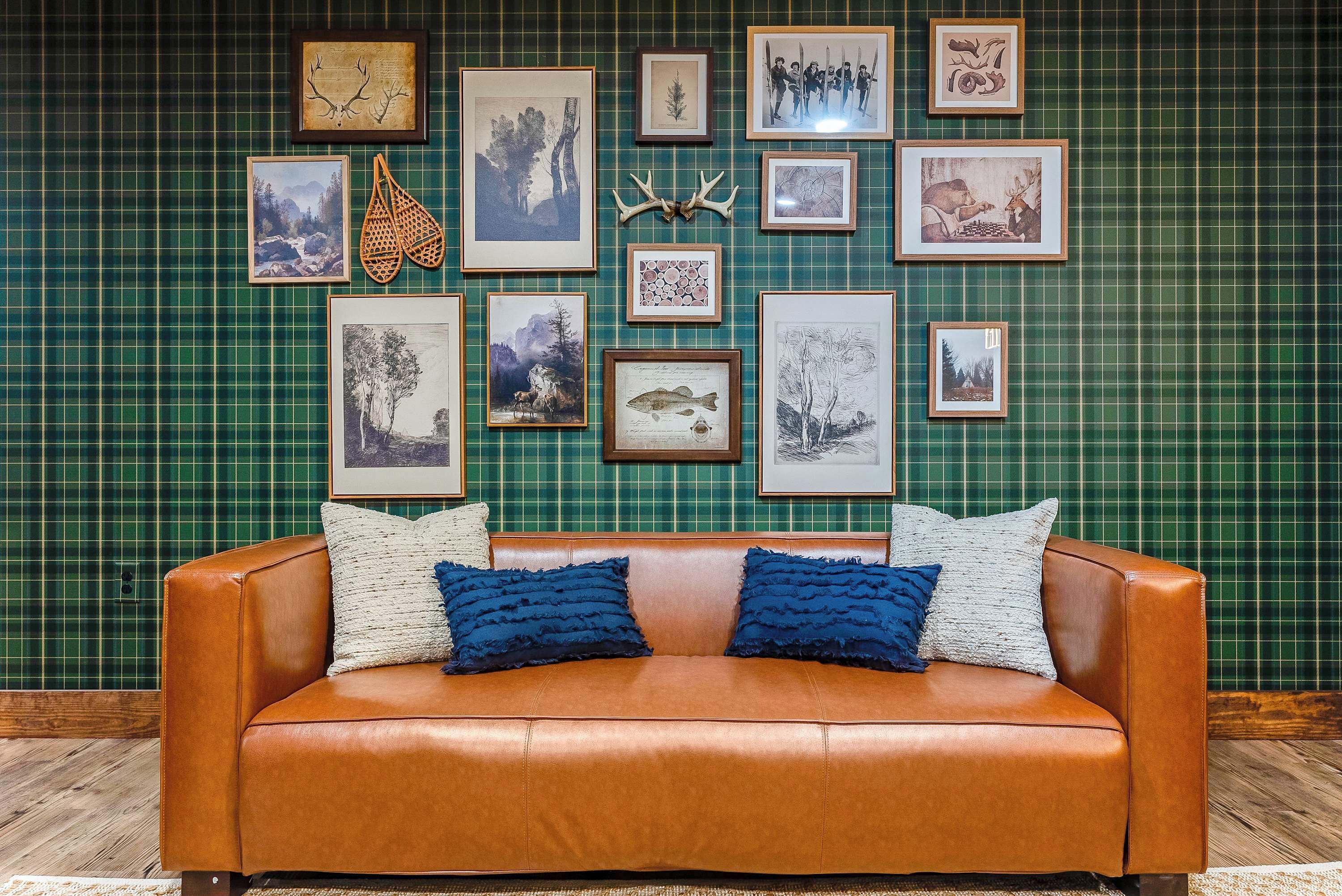 A brown leather sofa with two blue pillows against a green plaid wall with framed pictures.