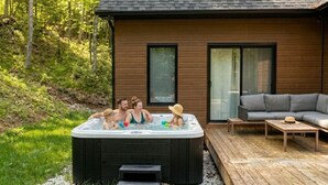 Un jacuzzi où se trouvent des personnes, une terrasse en bois, un canapé et une maison avec de grandes fenêtres.