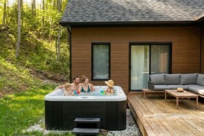 Ein Whirlpool mit Personen, eine Holzterrasse, eine Sitzecke und ein Haus mit großen Fenstern.