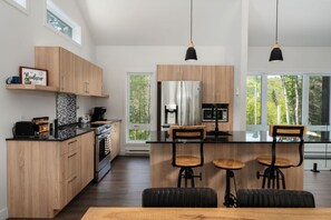 A modern kitchen with wooden cabinets, a central island with stools, a stainless steel refrigerator, and a view of trees through large windows.