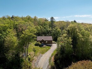 Una casa de un piso con techo inclinado, rodeada de un bosque frondoso, accesible por un camino de grava.
