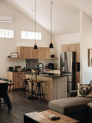 A modern kitchen with wooden cabinets, a central island, bar stools, and a dining area with a sofa and table.
