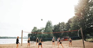 Un match de volleyball de plage se déroulant sur un terrain sablonneux, entouré d’arbres et bordé par une étendue d’eau.