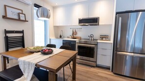 A modern kitchen with stainless steel appliances, a dining table with a bowl of fruit, and a pendant light. - Private Spa| Hiking| Skiing| Mini Chalet| Assinie (Brownsburg)