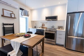A modern kitchen with stainless steel appliances, a dining table with a bowl of fruit, and a pendant light. - Private Spa| Hiking| Skiing| Mini Chalet| Assinie (Brownsburg)
