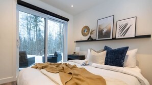 A modern bedroom with a large bed, a nightstand, a window with a view of trees, and framed artwork on the wall. - Private Spa| Hiking| Skiing| Mini Chalet| Assinie (Brownsburg)